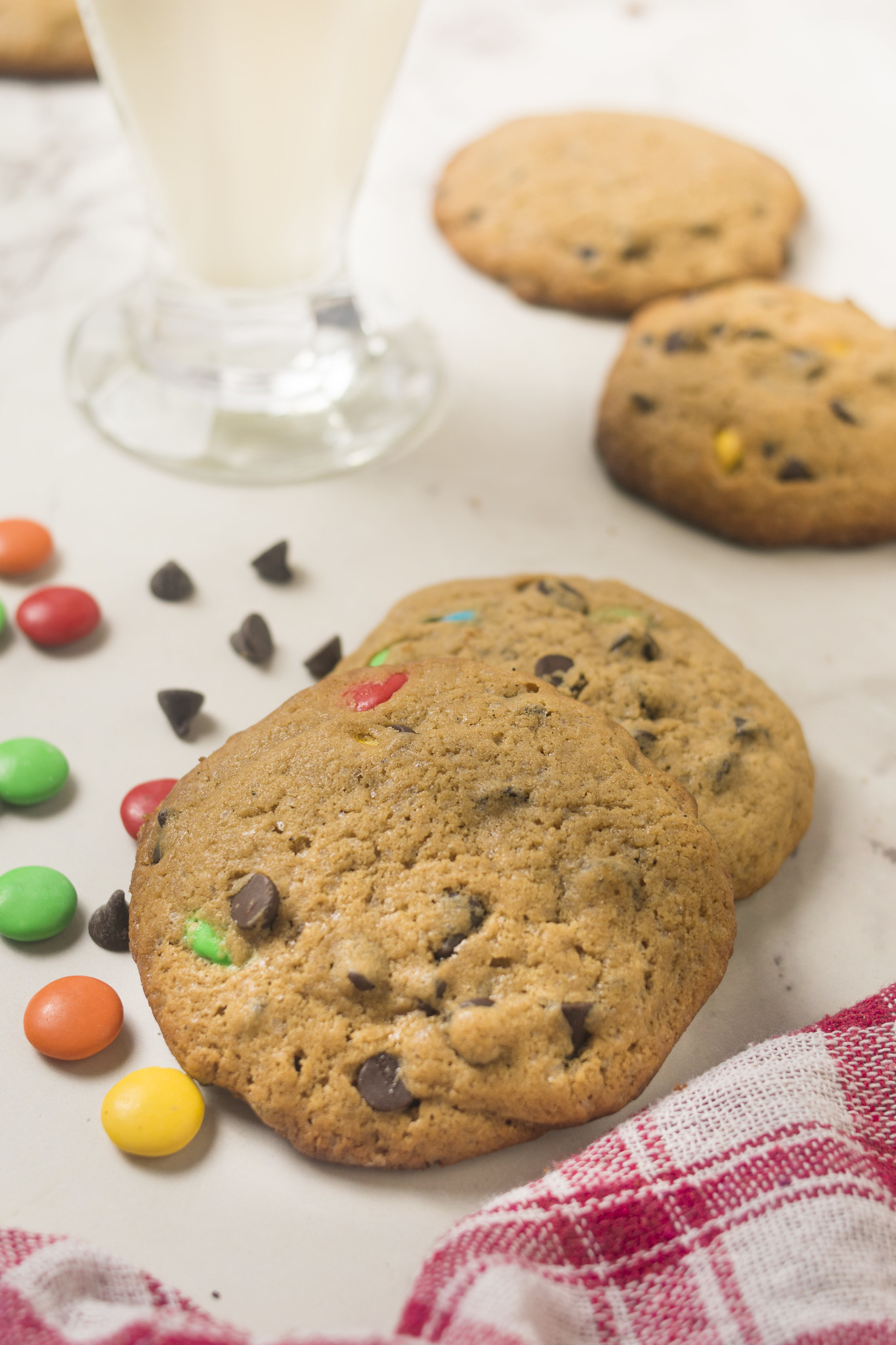 Browned butter chocolate chip cookies with colourful M&Ms, chocolate chips, a glass of milk and red gingham cloth.